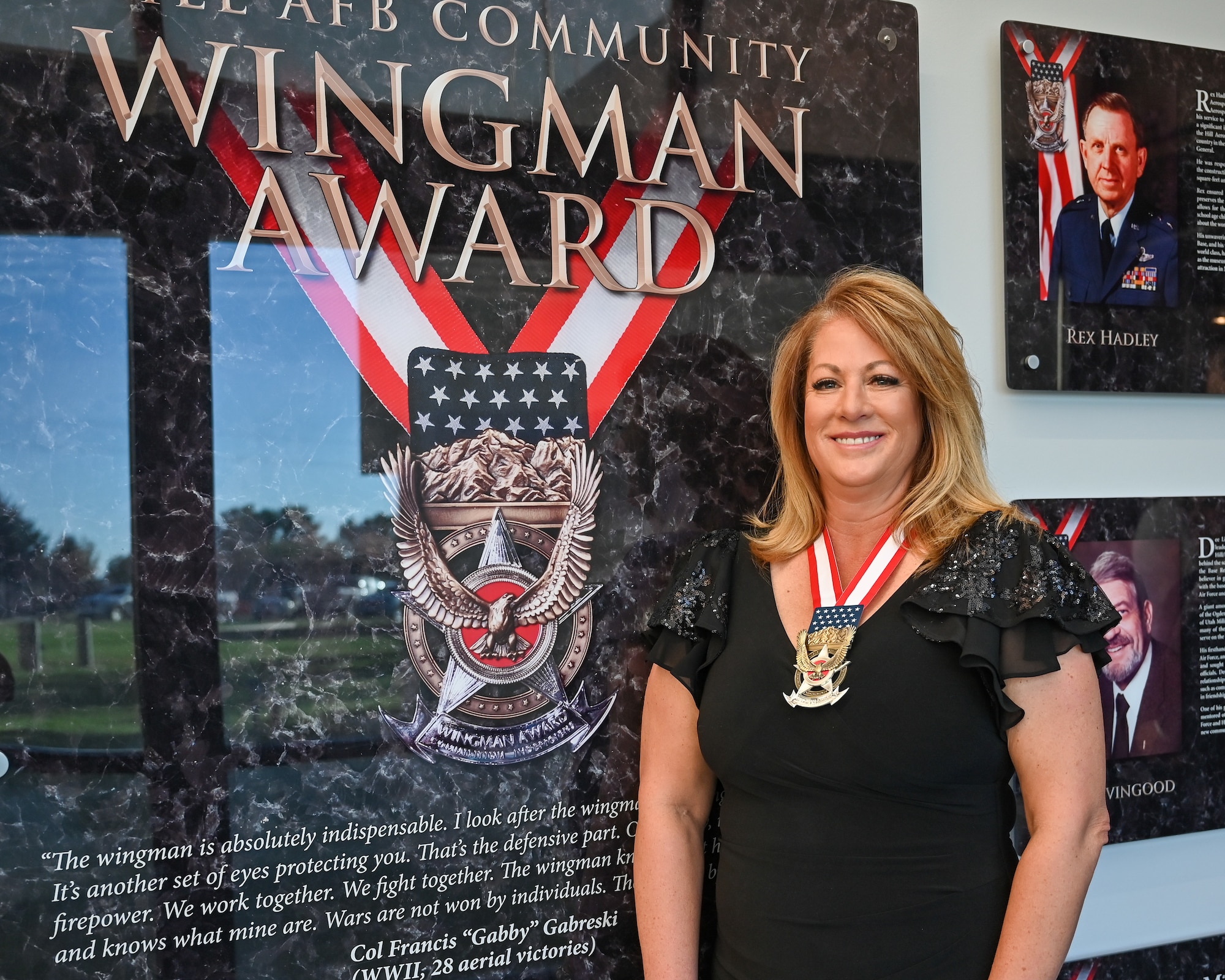 Lacy Bizios stands next to the Community Wingman Award wall display wearing her Community Wingman Award medallion.
