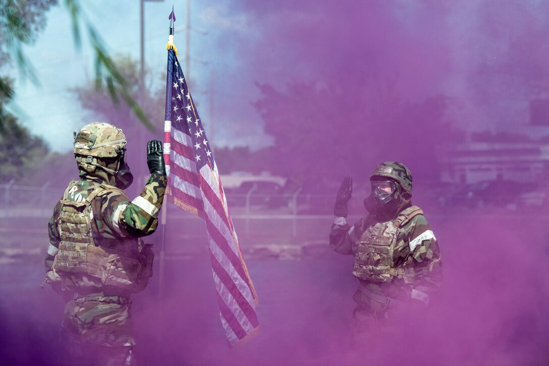 Two airmen in protective gear face each other with right hands raised surrounded by purple smoke.