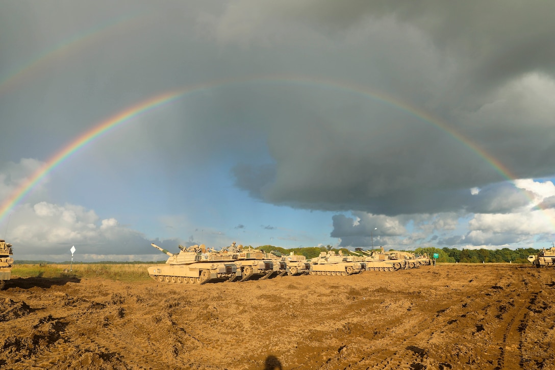 Tanks line up in the dirt under two rainbows.