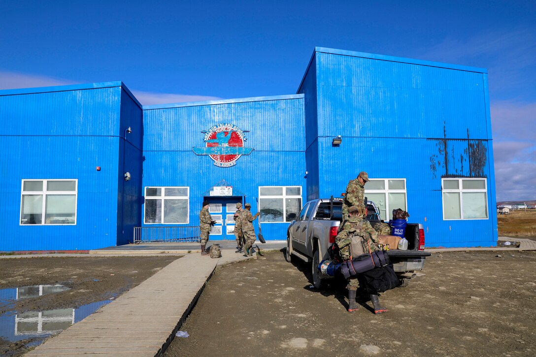 Service members unload gear from a pickup truck parked outside a blue building.