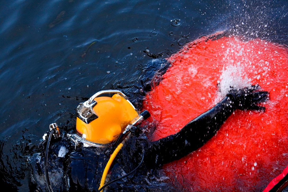 A sailor wearing dive gear holds onto a red ball in waters.