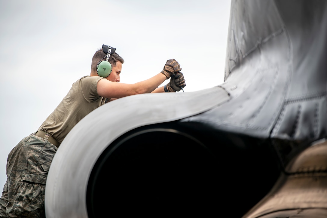 An airman wearing headphones and work gloves uses a tool to tend to an aircraft.