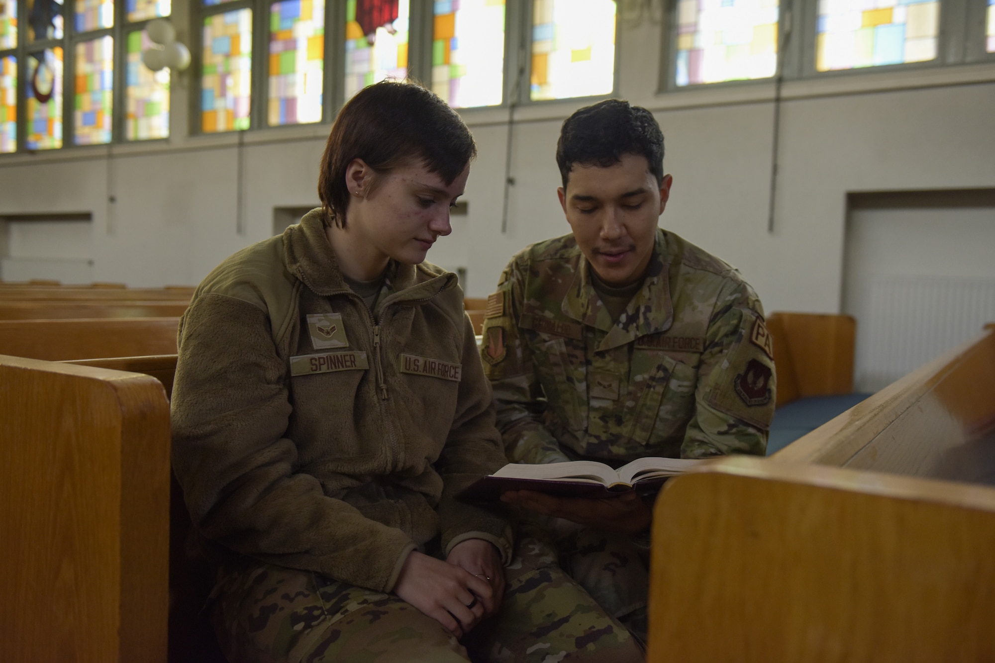 A woman, on the left, and a man, on the right, read a hymnal together while sitting on pews in a chapel.