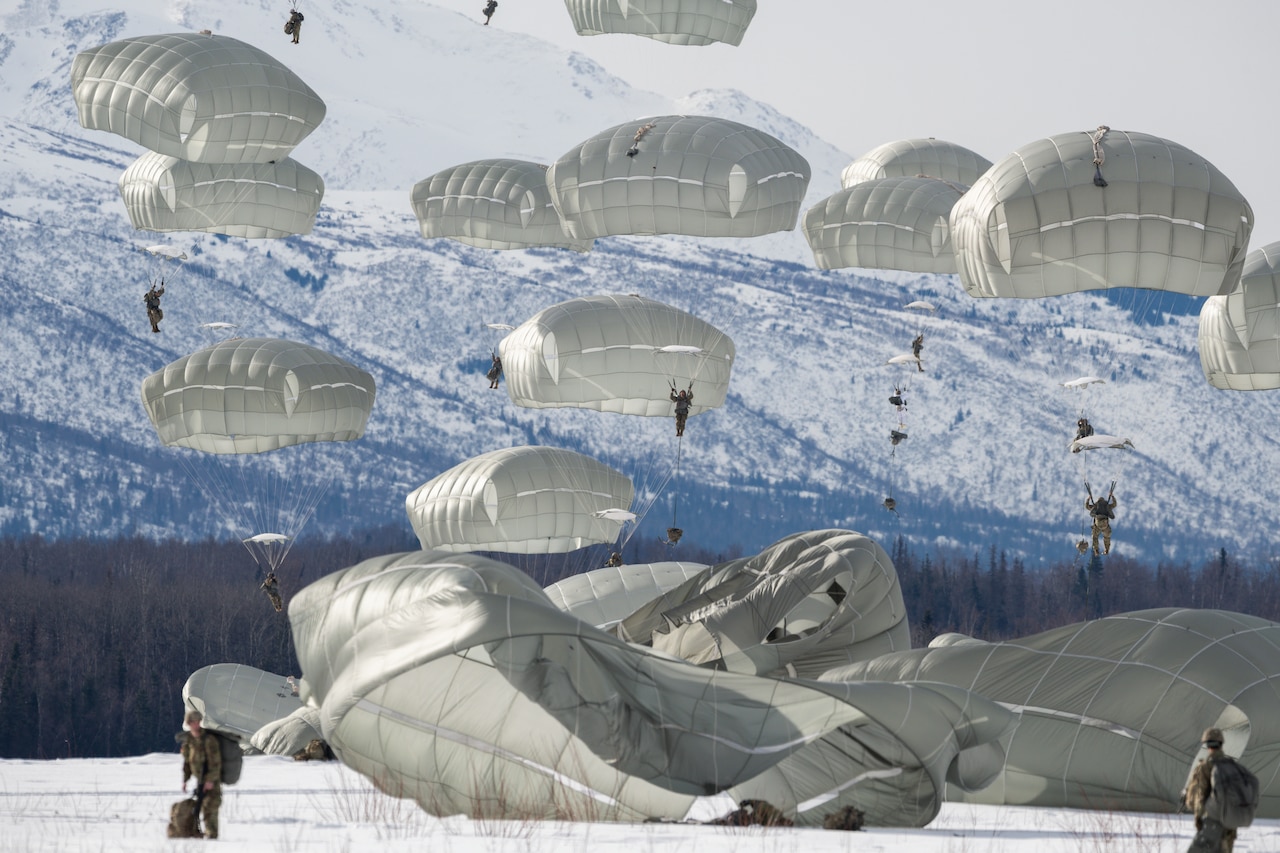 More than a dozen parachutes dot the sky as paratroopers land in a snow-covered, mountainous area.