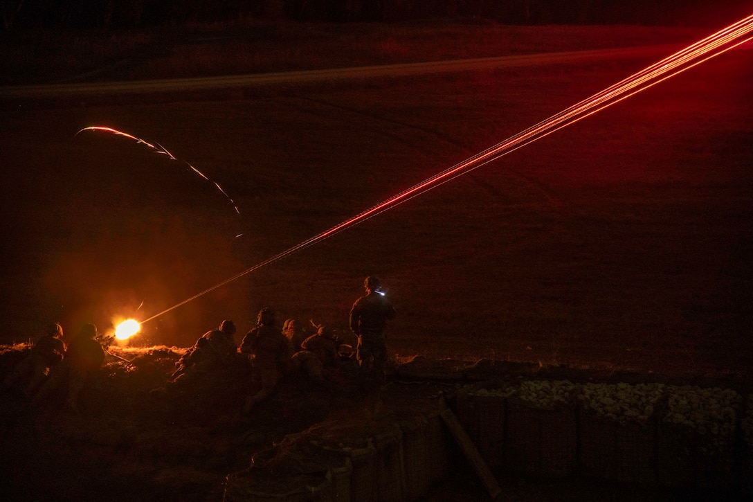 Soldiers laying on the ground fire weapons in the dark illuminated by orange light.