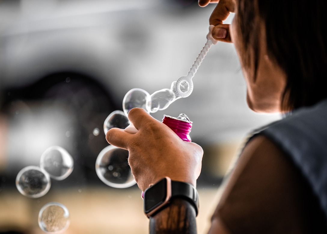 A woman with brown hair and a smart watch blows bubbles. On her arm is a tattoo.
