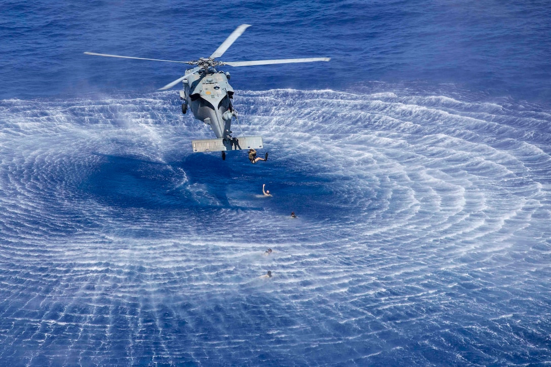 A sailor hangs from a rope from an airborne helicopter over water as others swim underneath.