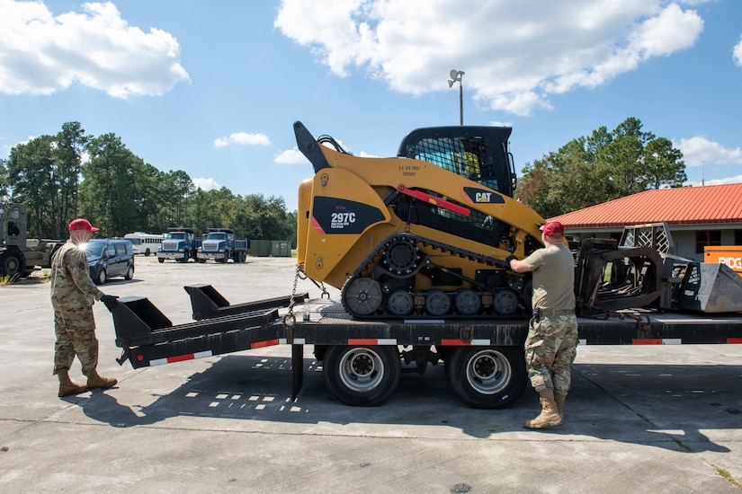 Service members stand near a trailer that carries a construction vehicle.