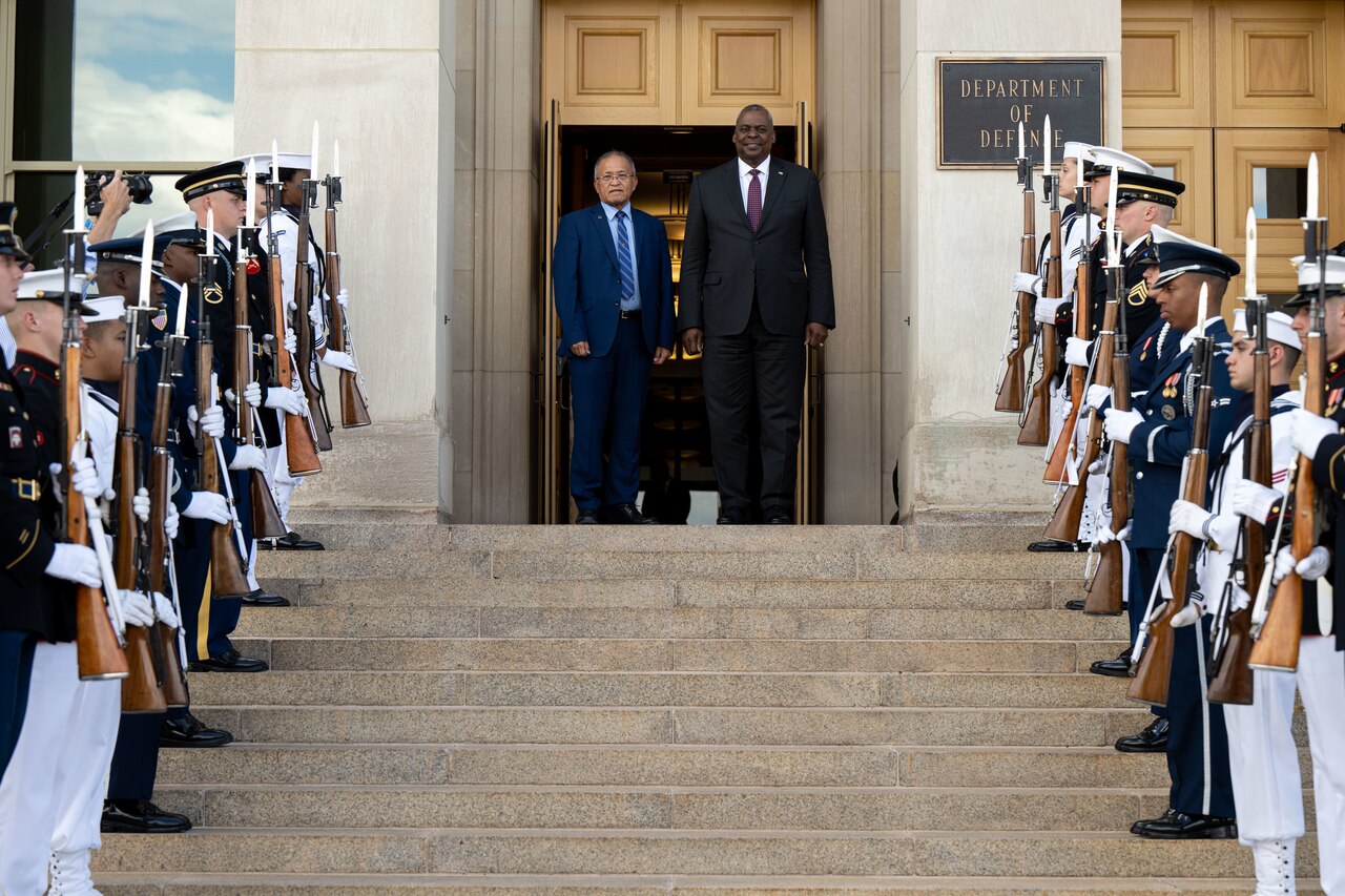 Two men stand in a doorway overlooking an honor guard. The plaque next to the door indicates that they are at the Department of Defense.