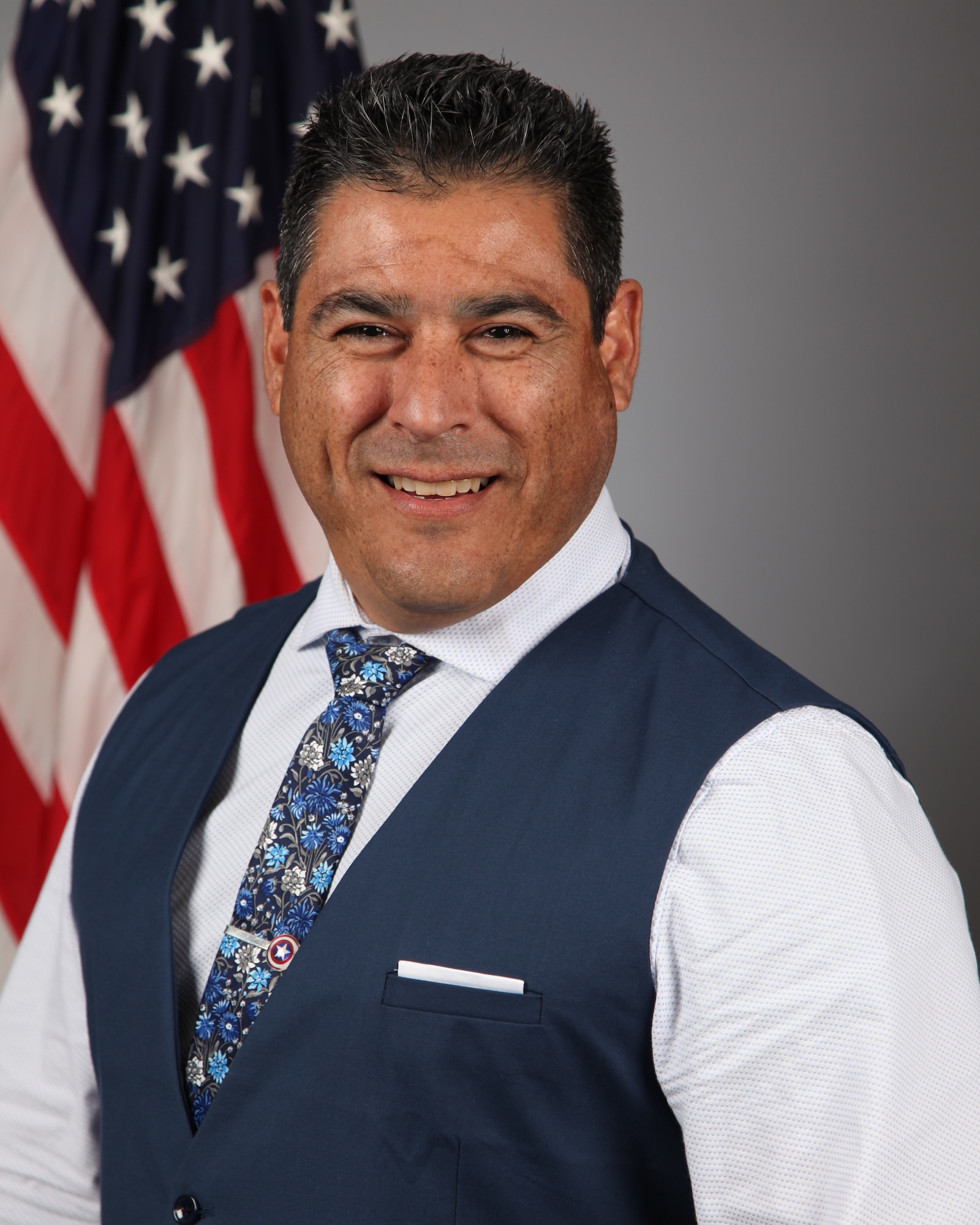 A man in a suit stands in front of a flag