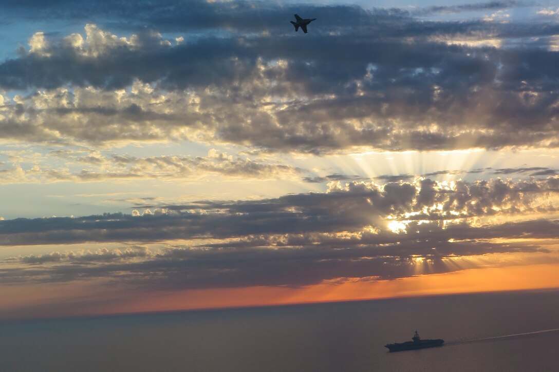 An aircraft flies above a ship amid a dramatic blue and pink sun-streaked sky.