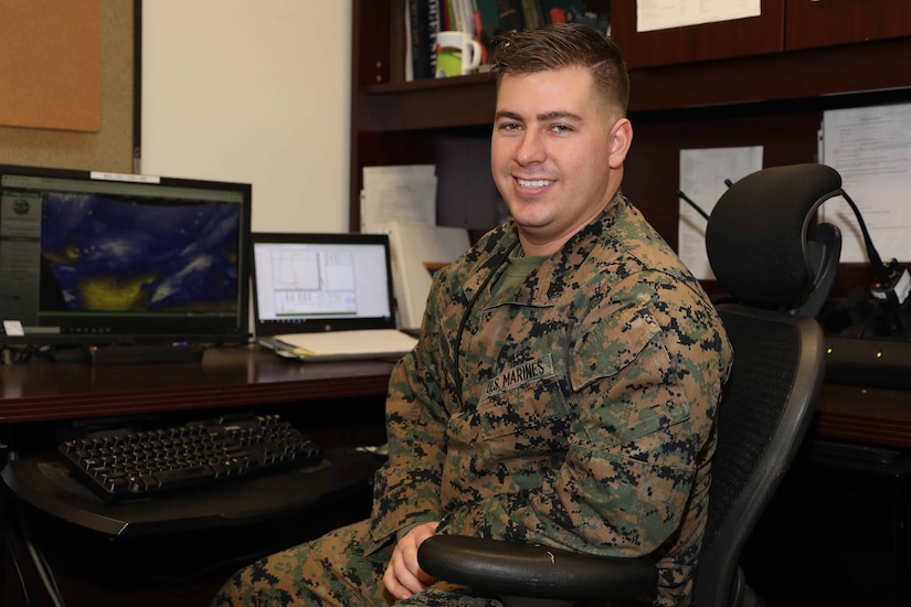A man in combat uniform smiles in front of a desk.