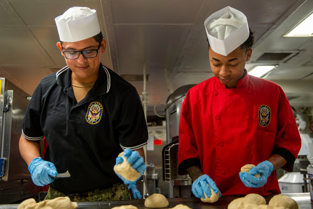 Two sailors knead dough in a kitchen aboard a ship.