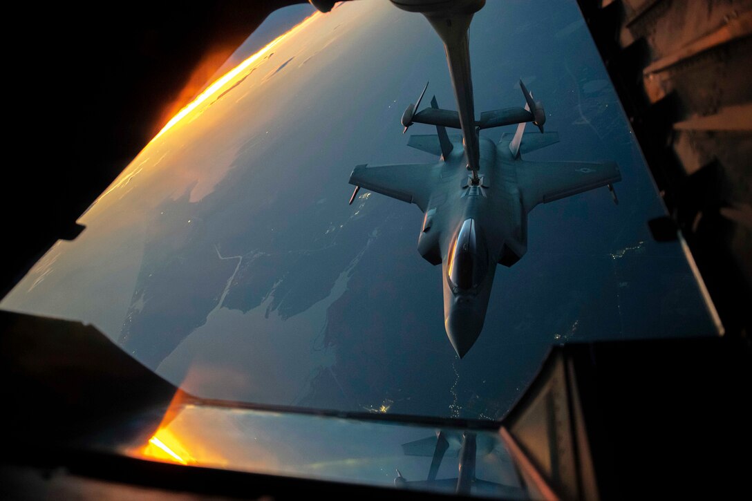 An aircraft receives fuel from another aircraft while flying under a sunlit sky.