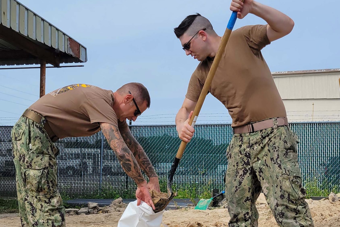 A sailor shovels sand into a bag held by another sailor.