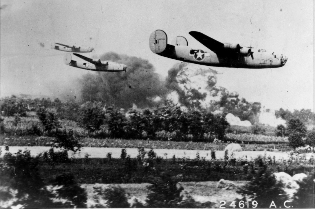 Three bomber aircraft fly low over land. Large plumes of smoke and fire are visible in the background.