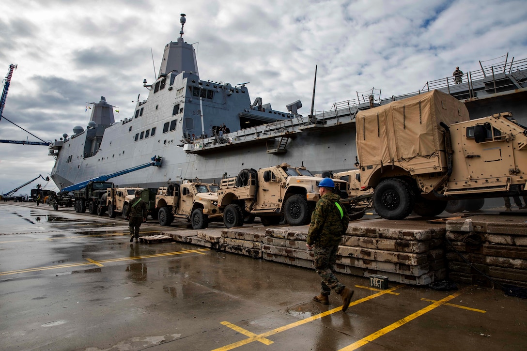 Two Marines walk by a line of military vehicles aside a docked ship.