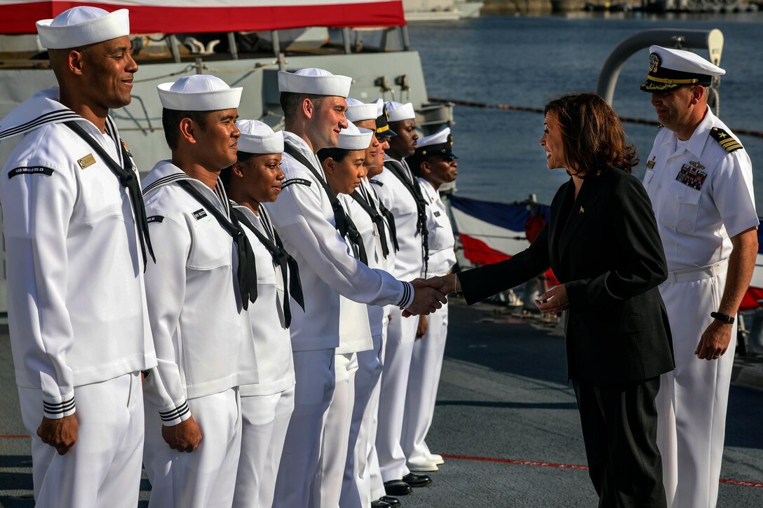 Vice President Kamala Harris shakes hands with sailors standing in a row on a ship.