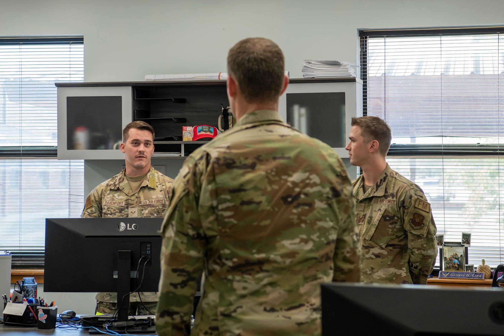 Two Airmen stand behind desks with the 7th Bomb Wing commander speaking to them.