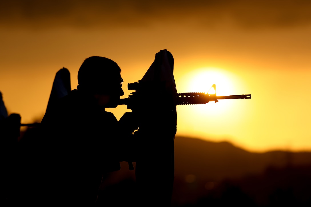 A Marine aims a weapon under a sunlit sky.