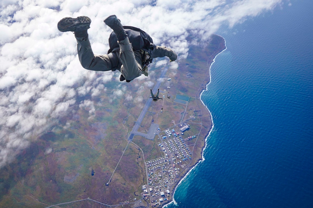 Sailors free fall wearing parachute packs over the coast of a city.