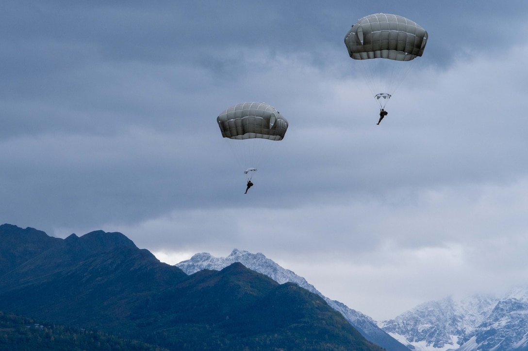 Two parachutists descend to the ground with mountains behind them.