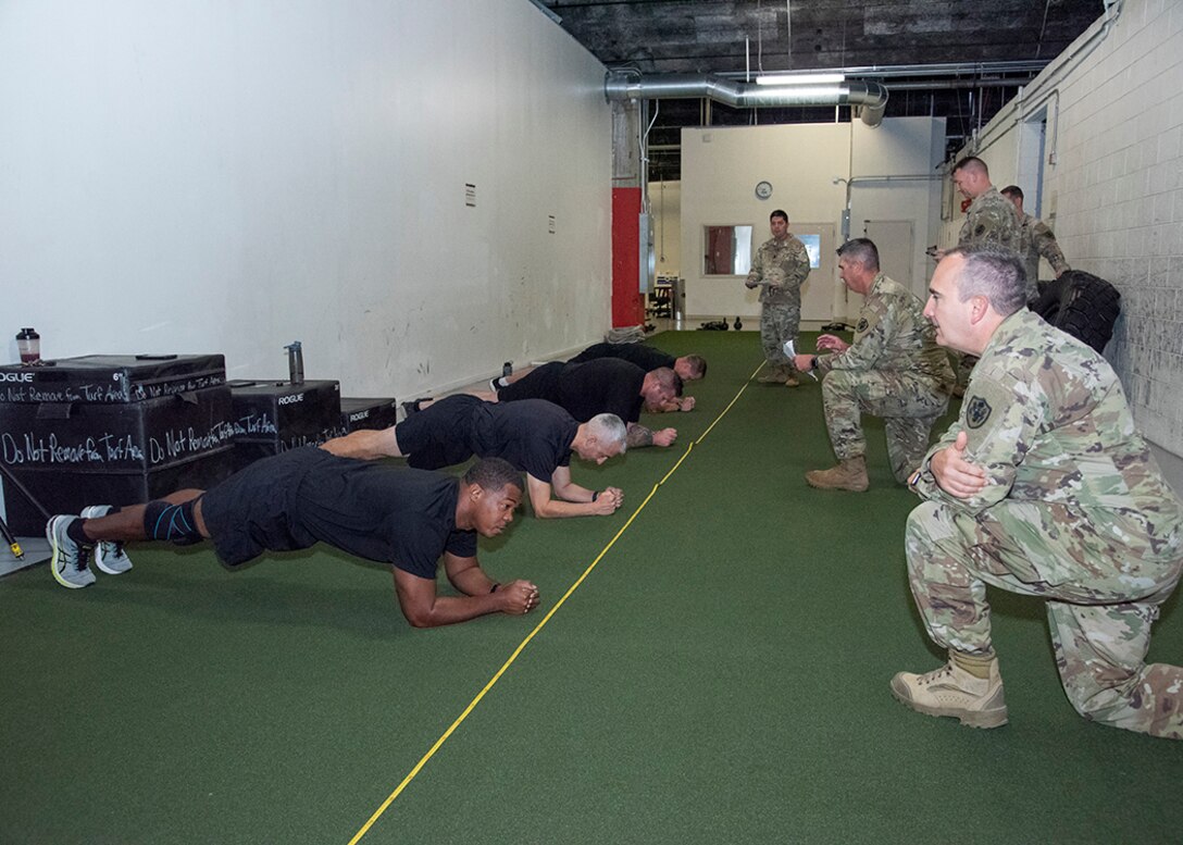 Four men perform a plank. They are dressed in black fitness gear. Other people in Army OCP uniforms observe them.