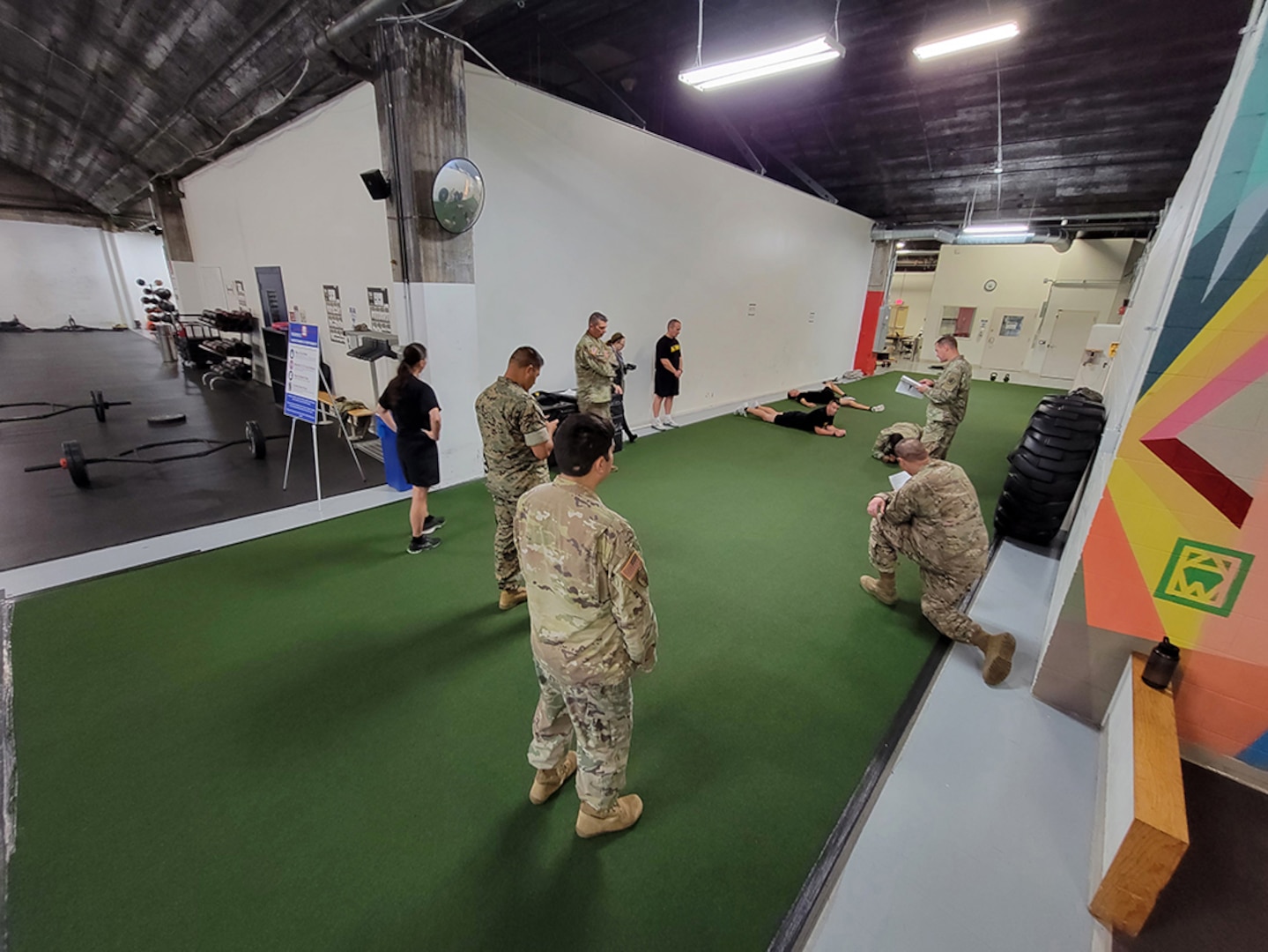 People in Army OCP uniforms and black fitness gear listen to instructions inside a fitness center.