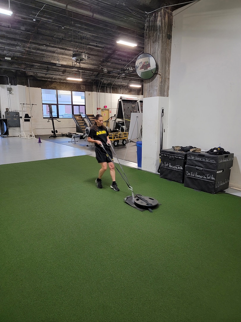 A woman in black fitness gear drags a weighted sled inside a fitness center.