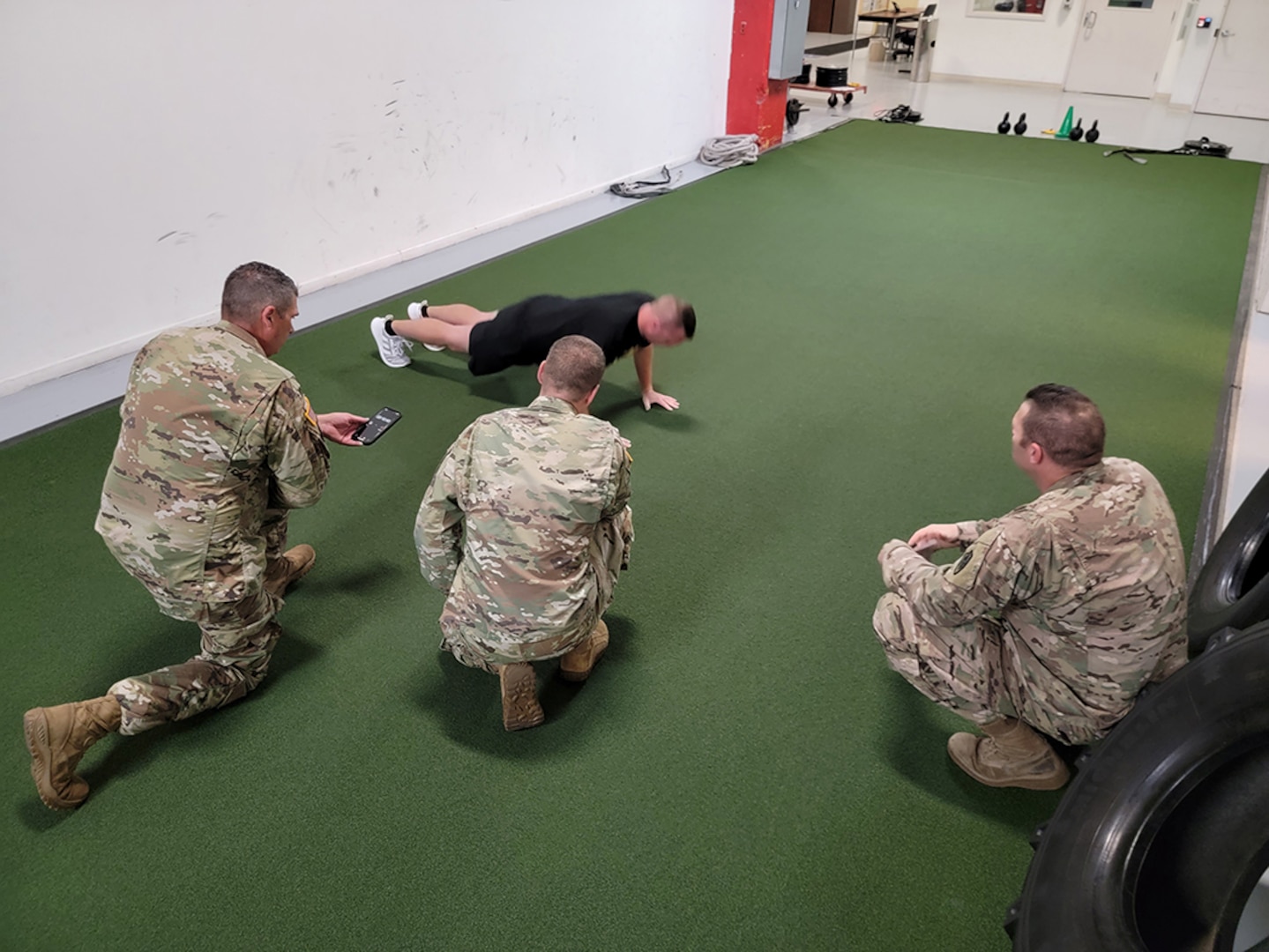 People in Army OCP uniforms watch another person in black fitness gear do a push-up.