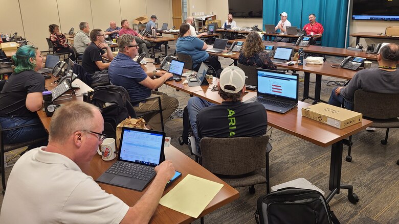People sit at rows of long, wooden tables in a conference room, all facing toward two instructors training the group to take calls at a call center.