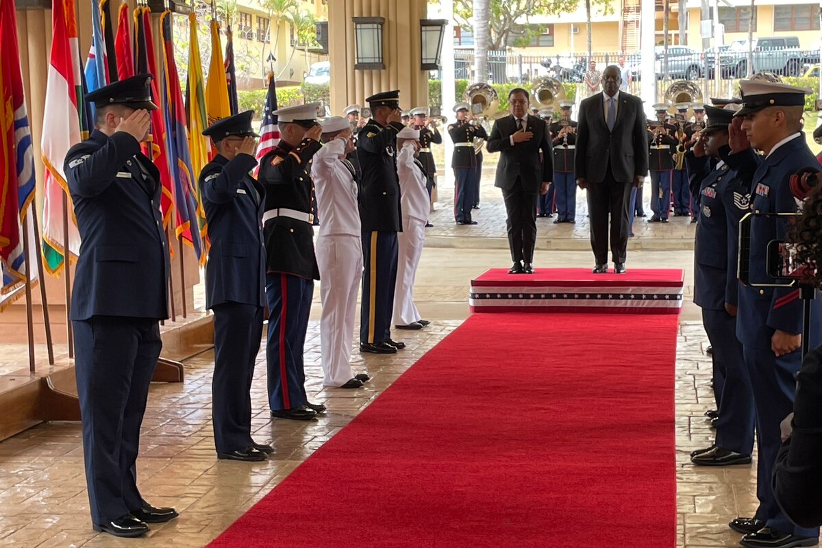 Secretary of Defense Lloyd J. Austin III and another official stand at one end of a red carpet, along which service members are saluting.