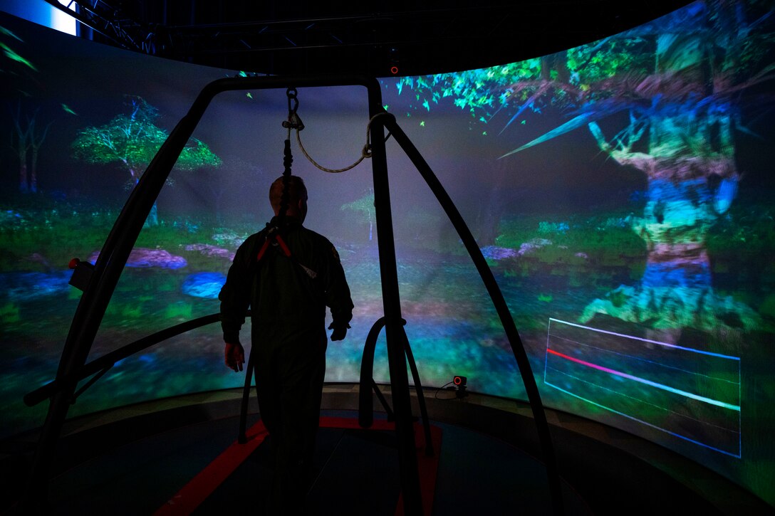An airman walks on a treadmill-type device facing a large curved screen with a virtual nature scene.