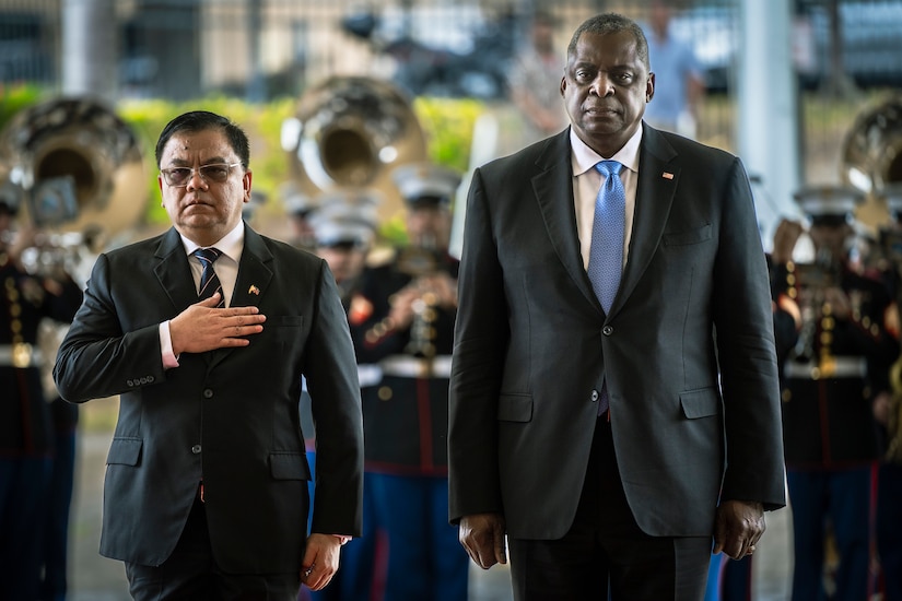 Secretary of Defense Lloyd J. Austin III stands next to another official outside, with military band in background.