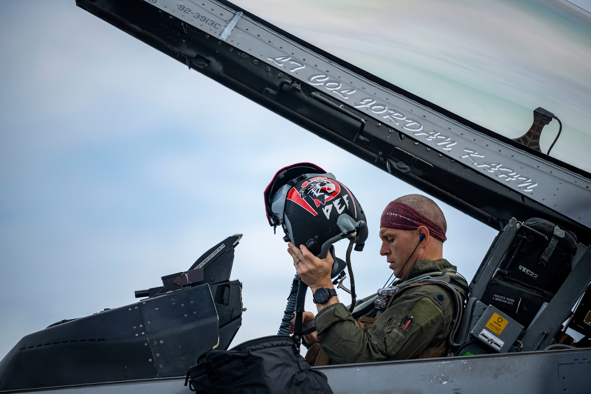 A military member in uniform puts on his decorated helmet in the cockpit of a jet.