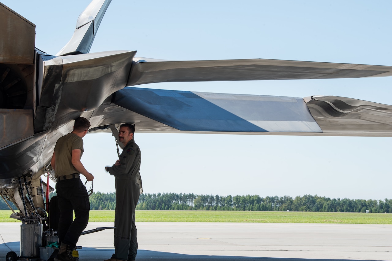 Two service members inspect underneath the wing of an airplane.