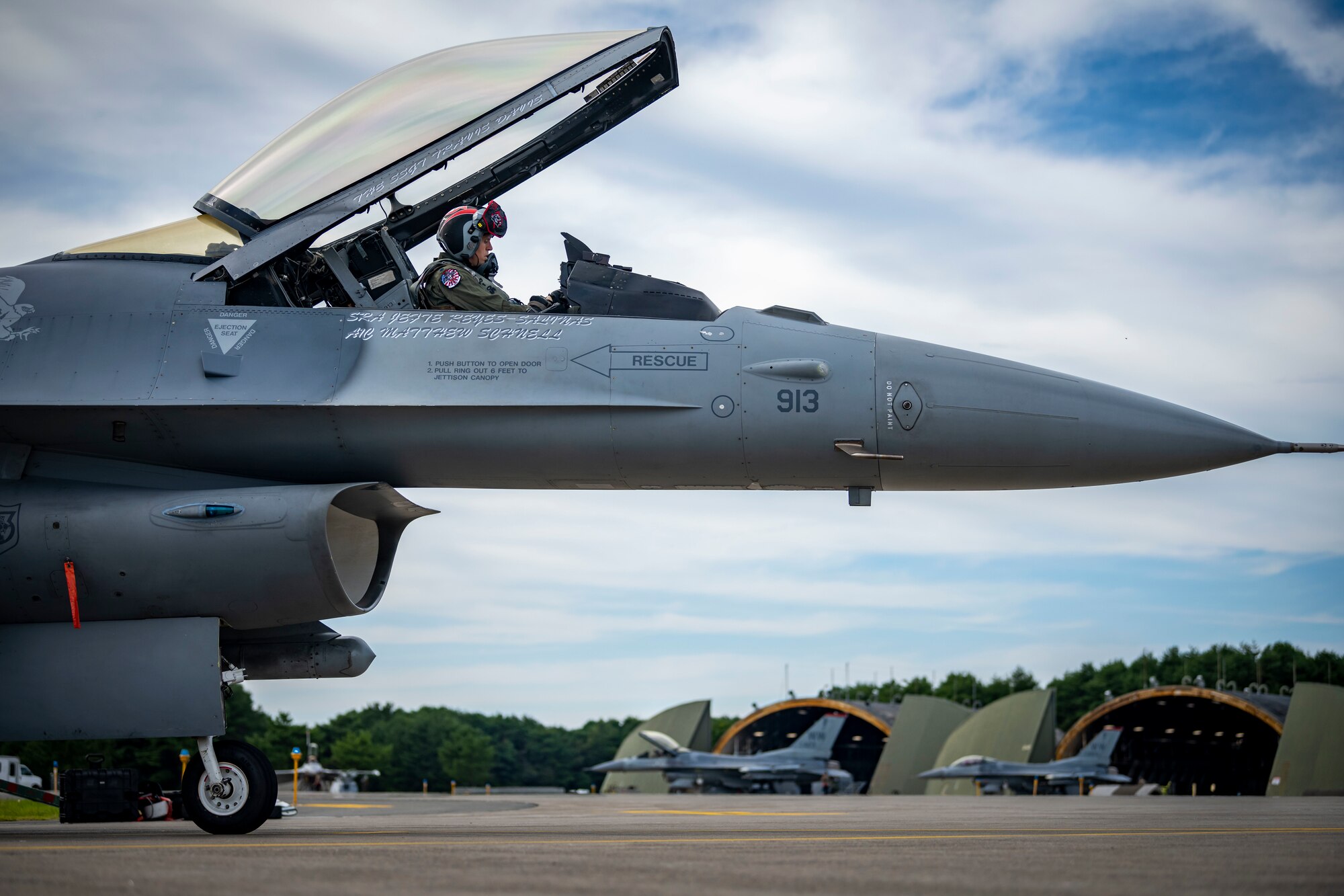 A military member in uniform straps in the cockpit of a jet.
