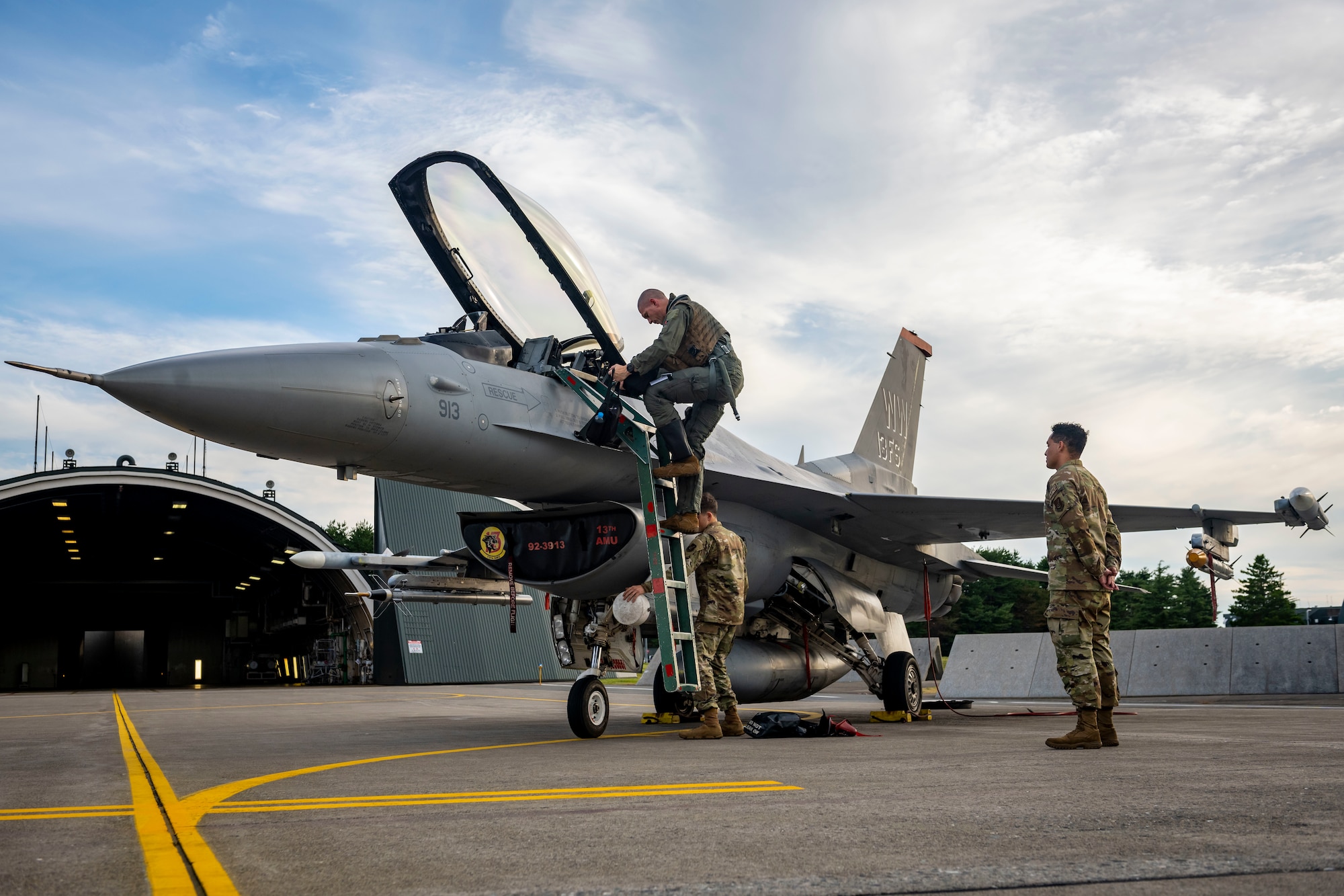 Two military members in uniform wait as a pilot climbs down the ladder of a jet.