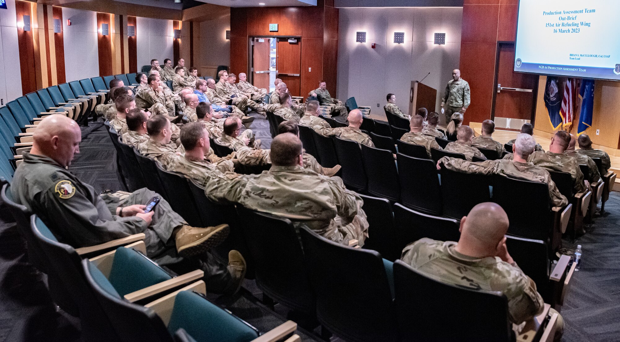 A big group of military members sit in a room for a briefing.