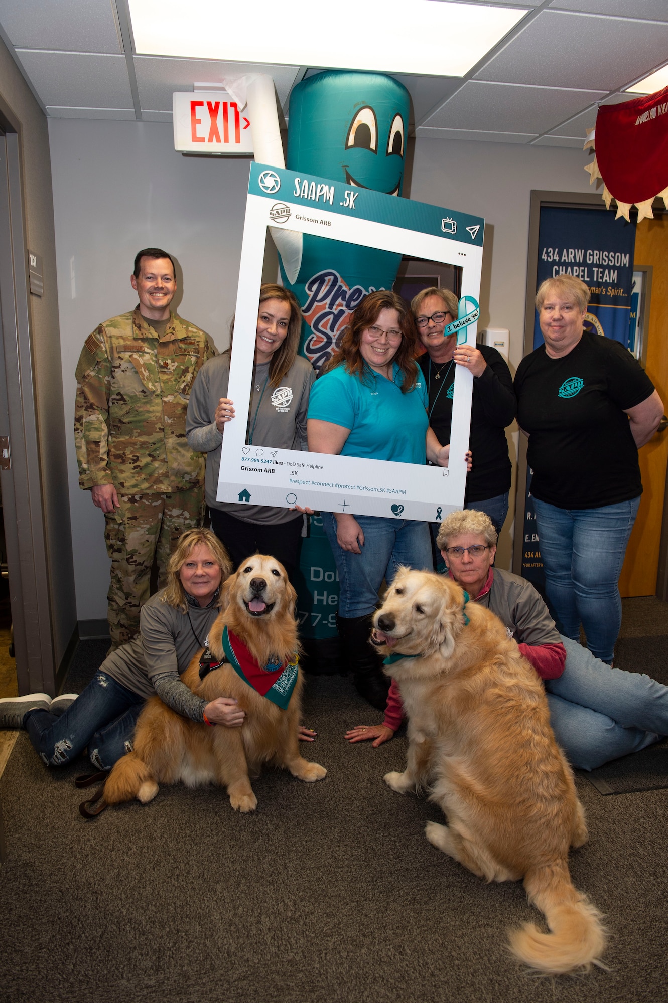 Group photo of sexual assault and prevention team and other volunteers posed with photo prop, and two golden retriever dogs in foreground.