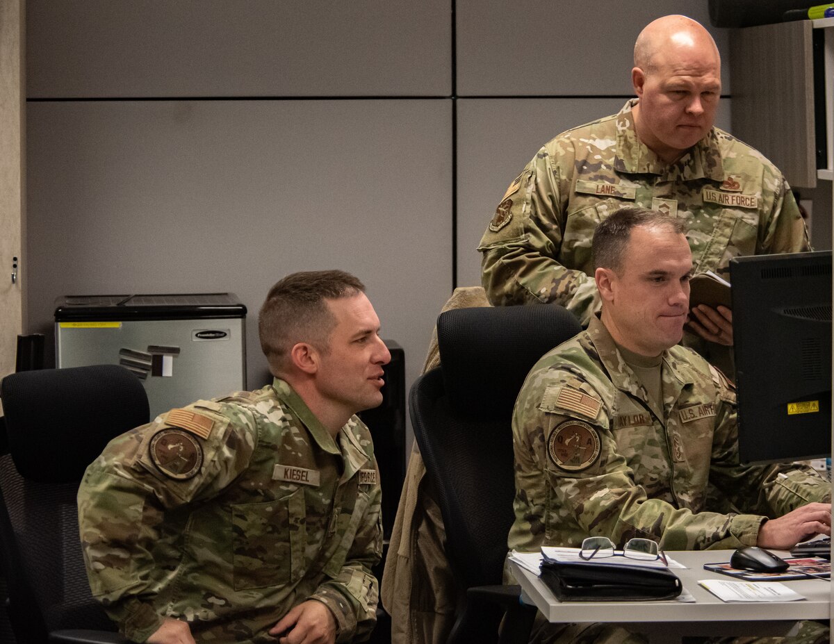 Three military members sit in a room around a computer.