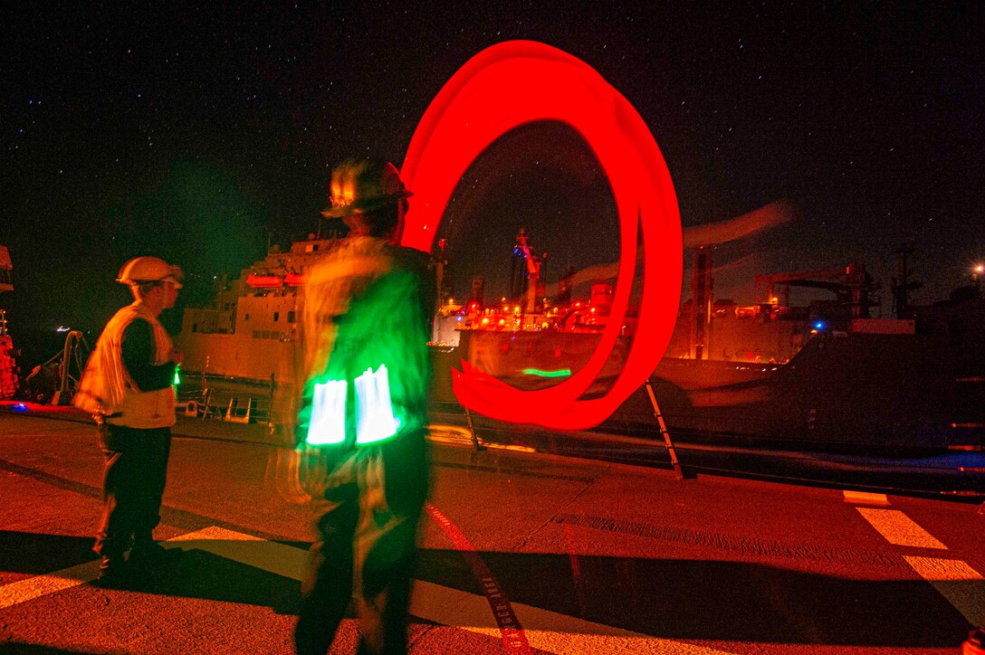 A sailor uses a red light to signal toward a ship as another sailor stands beside.