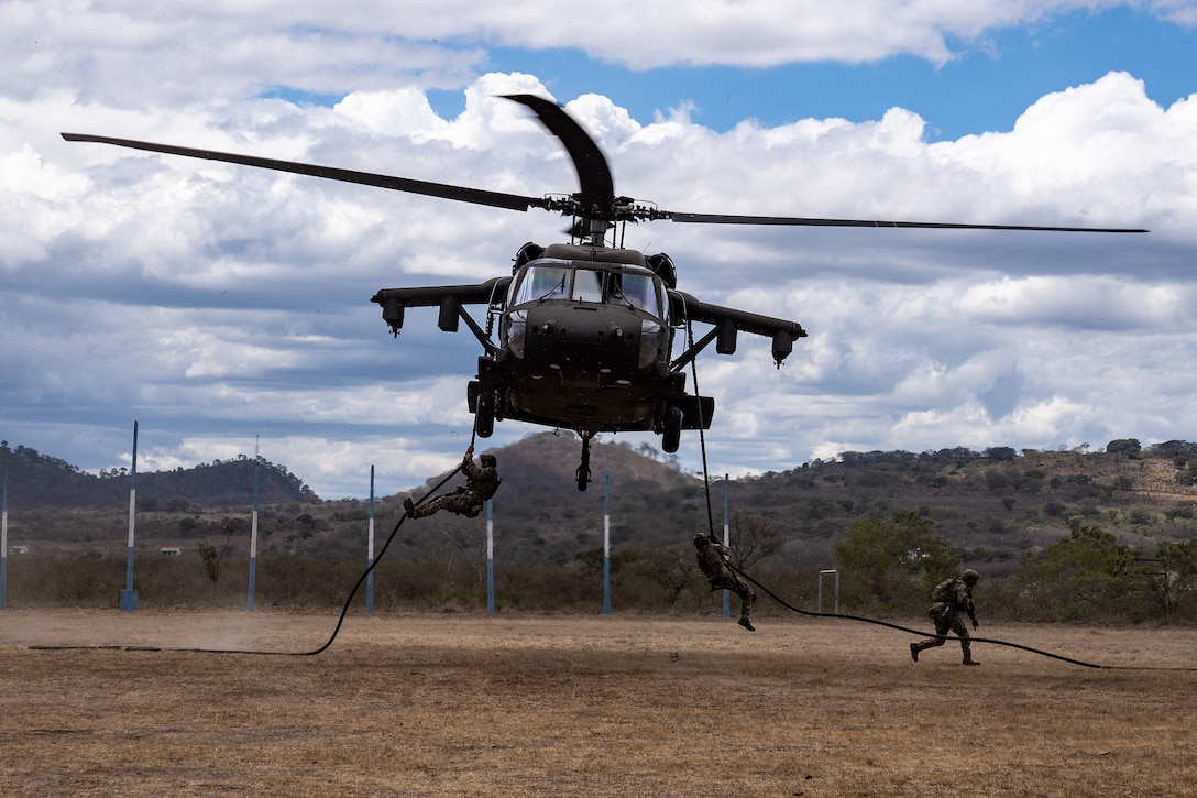 Soldiers hold onto ropes from the sides of a military helicopter as it hovers over land.