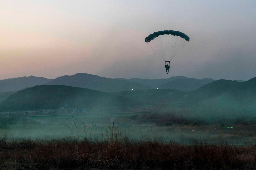 A Marine descends with a parachute over mountains surrounded by fog.