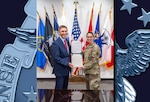 A grey haired man in a blue suit holds a proclamation with a dark haired woman in Army OCPs with a flag display behind them and a shiny table in front of them. The paper is reflecting off the table.
