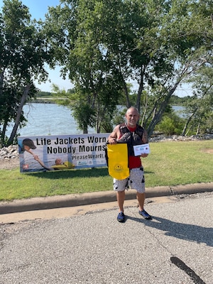 Man wearing red life jacket holding yellow dry bag and ticket saying "I got caught wearing my life jacket" stands in front of a sign stating "life jackets worn... nobody mourns" with two people in a red and yellow life jacket reach for one another.