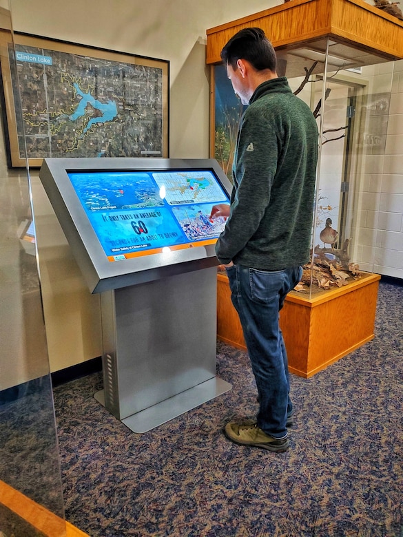 Man wearing blue jeans and a green zip-up jacket standing next to the interpretive kiosk using his hands to interact with the touch screen on the kiosk.