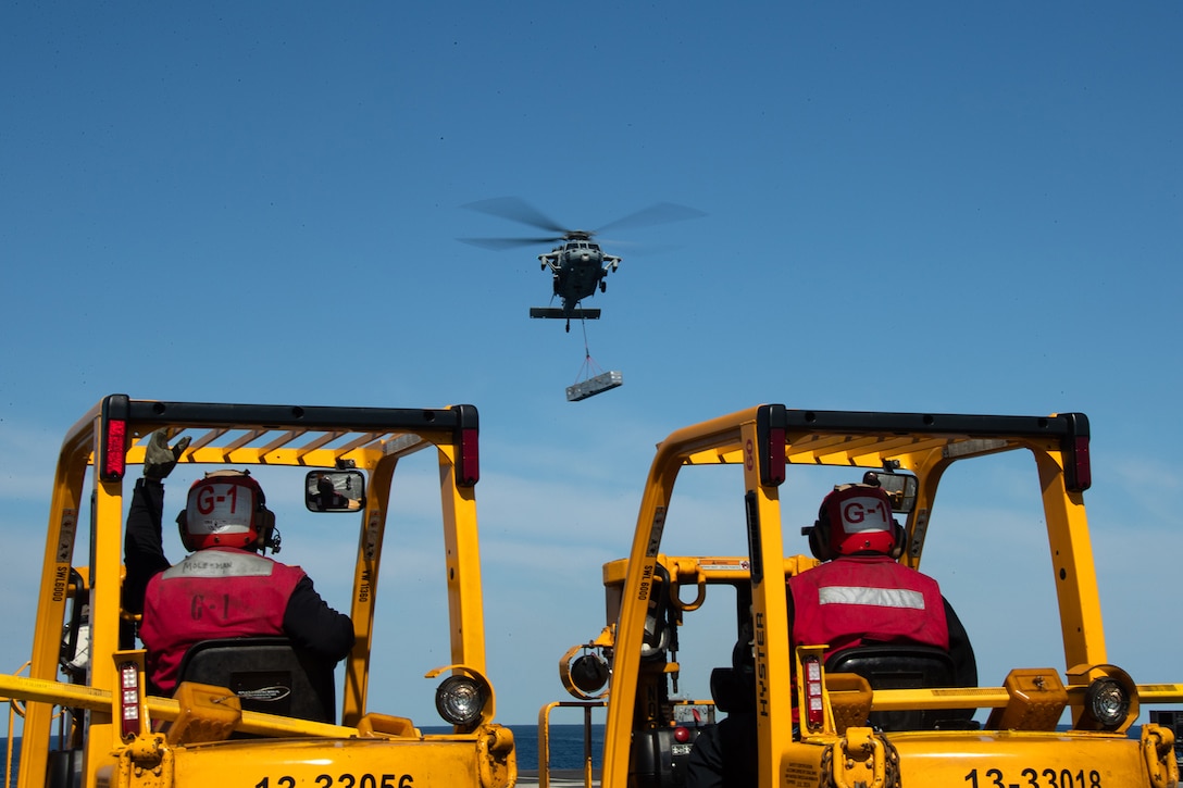 A Navy helicopter flies as two sailors operate machinery aboard the flight deck of a ship.