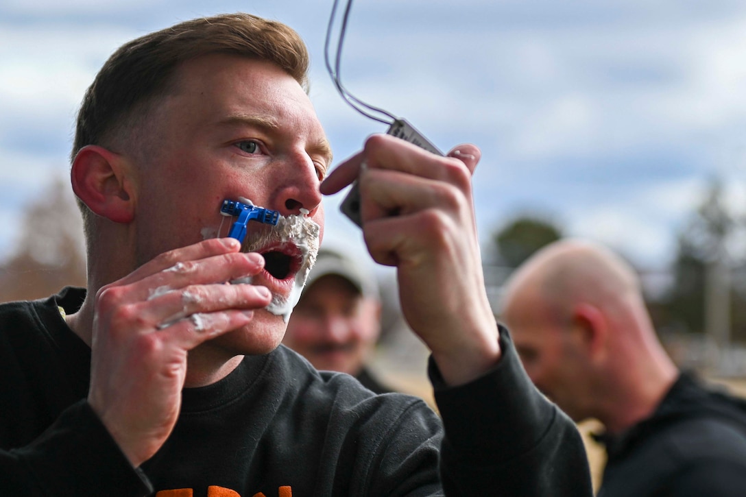 An airman holds up a small mirror while shaving his face with a razor.