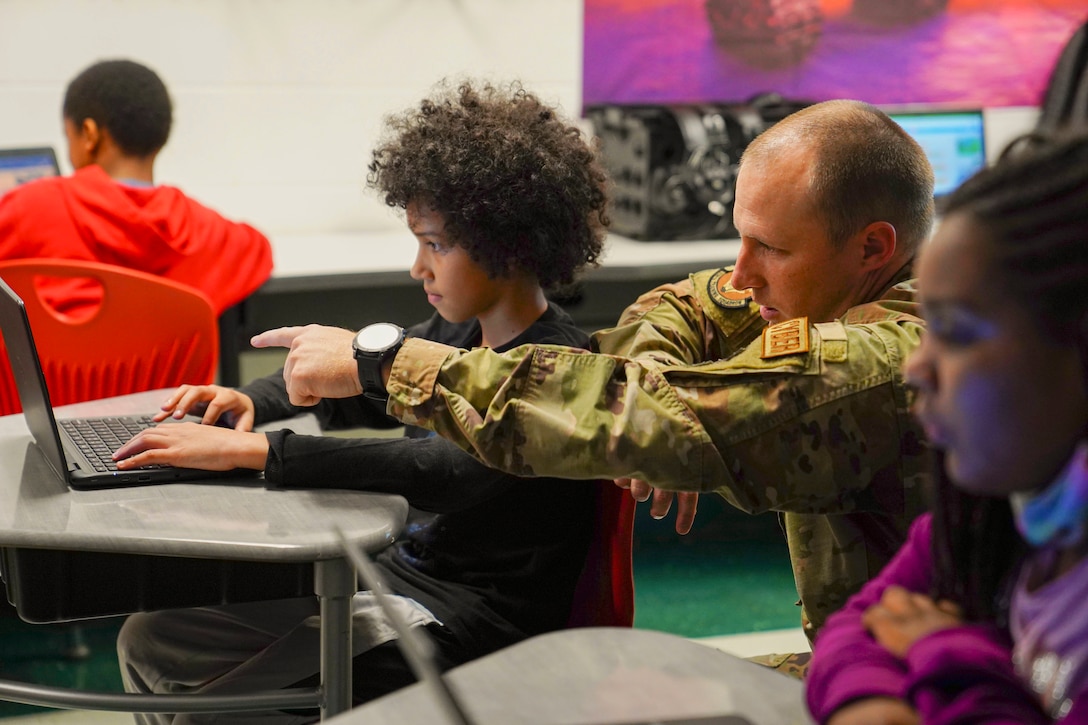 An airman kneels next to a student working a on a laptop in a classroom.